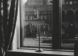The window looks out on the town hall, in front of which a Nazi banner hangs. The menorah belonged to Rabbi Akiva Posner, the rabbi of Kiel. 1931